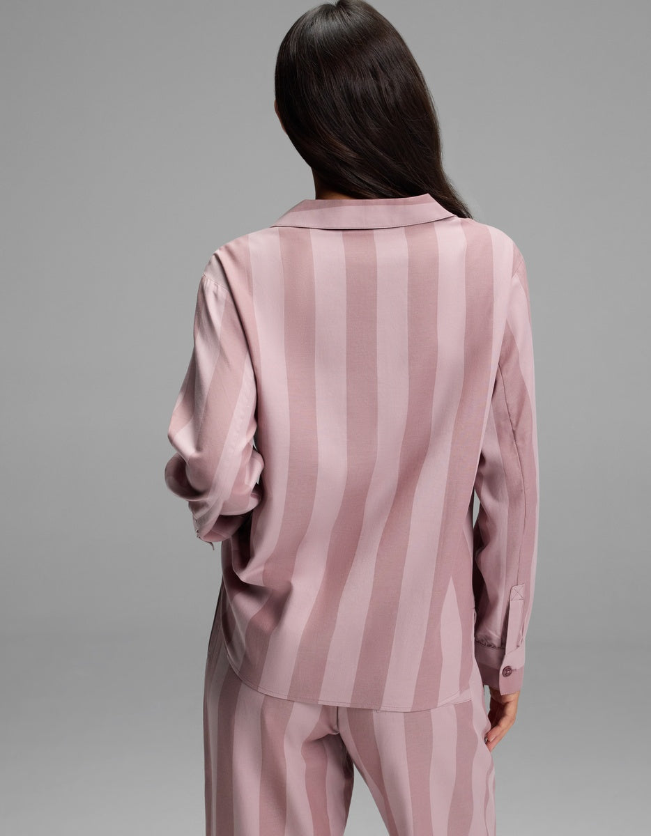 A woman with long dark hair is shown from the back, wearing the Cozy Earth Women’s Belmont Long Sleeve Pajama Top in a matching pink striped set, standing against a plain gray background.
