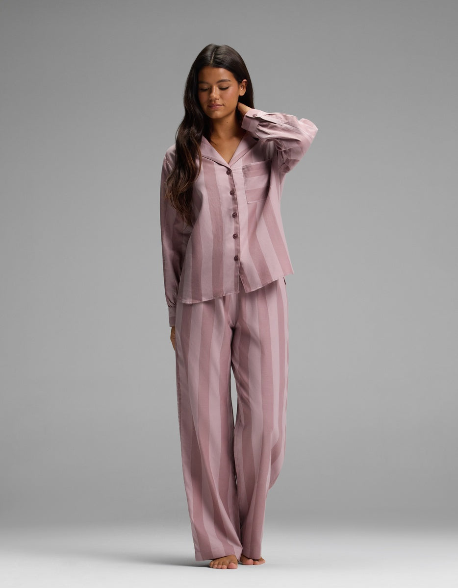 A woman stands barefoot on a gray background, wearing the Cozy Earth Women’s Belmont Long Sleeve Pajama Top in a loose mauve stripe pattern, paired with matching long pants. She has long dark hair and gently touches her neck with one hand.