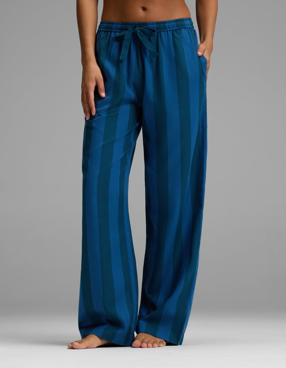 A person stands barefoot against a plain gray background wearing Cozy Earth’s Women’s Belmont Pajama Pant—a loose-fitting, blue striped style with a comfortable drawstring waist and hands relaxed by their sides.