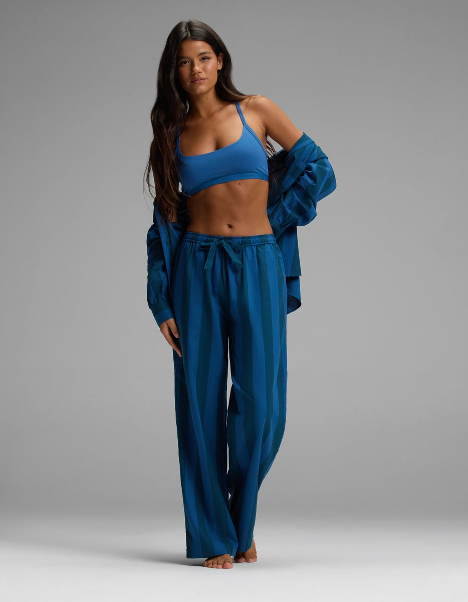 A woman with long dark hair wears Cozy Earth’s Women’s Belmont Pajama Pant, a matching blue sports bra, and an open blue shirt. She stands barefoot against a plain gray background, looking at the camera.