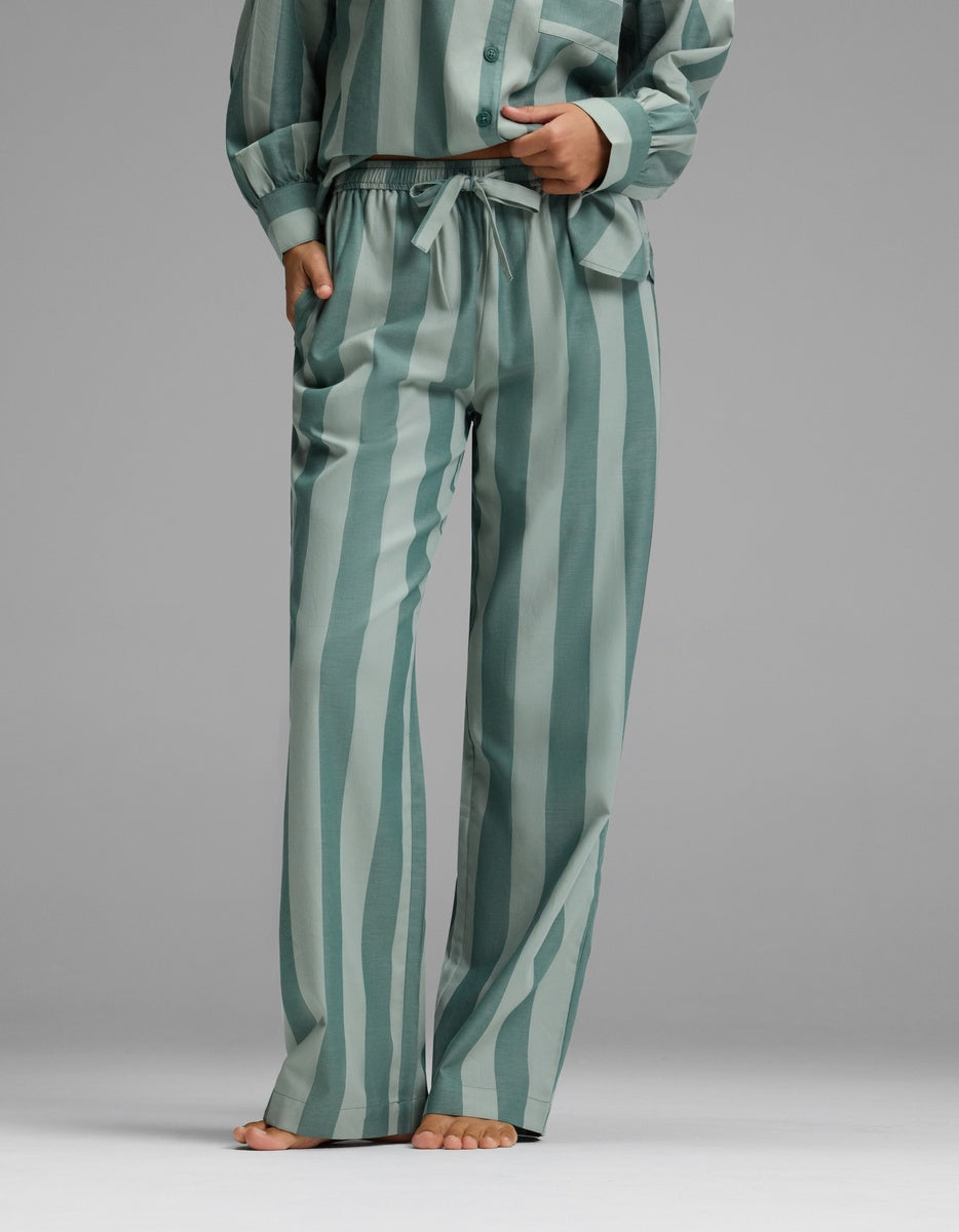 A woman is seen standing barefoot against a plain gray background, wearing Cozy Earth’s Women’s Belmont Pajama Pant in green with vertical stripes, paired with the matching long-sleeve top.