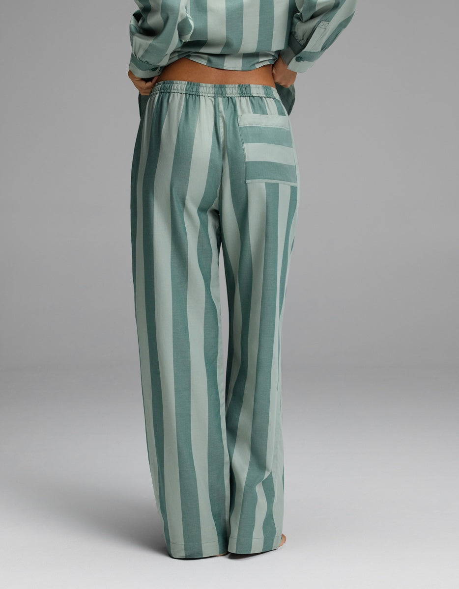 A person stands with their back to the camera, wearing the Cozy Earth Women’s Belmont Pajama Pant—a loose, high-waisted style in green and light green stripes—paired with a matching top. The pants feature a right-side pocket with horizontal stripes.