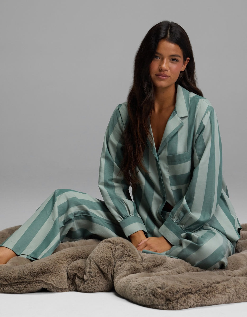 A woman with long dark hair sits on a furry blanket, looking relaxed as she models the Cozy Earth Women’s Belmont Pajama Pant against a plain gray background.