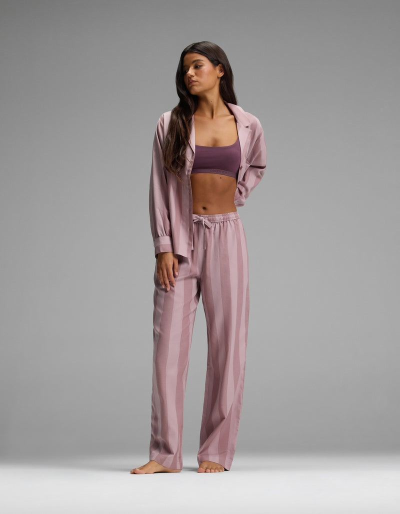 A woman stands barefoot against a plain gray background, wearing the Cozy Earth Women’s Belmont Pajama Pant in a pink striped design paired with a matching open shirt, revealing a purple sports bra underneath.