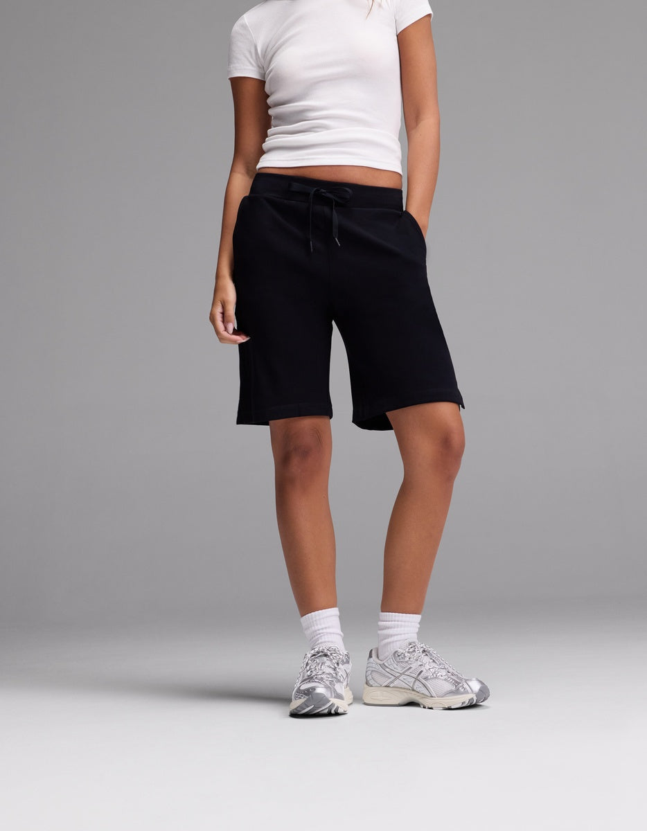 A person stands against a plain grey background wearing Cozy Earth Women’s Brushed Bamboo Bermuda Shorts, paired with a white fitted t-shirt, white socks, and white sneakers. Their hands are in their pockets and their face is not visible.