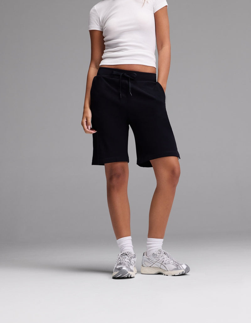 A person stands against a plain grey background wearing Cozy Earth Women’s Brushed Bamboo Bermuda Shorts, paired with a white fitted t-shirt, white socks, and white sneakers. Their hands are in their pockets and their face is not visible.