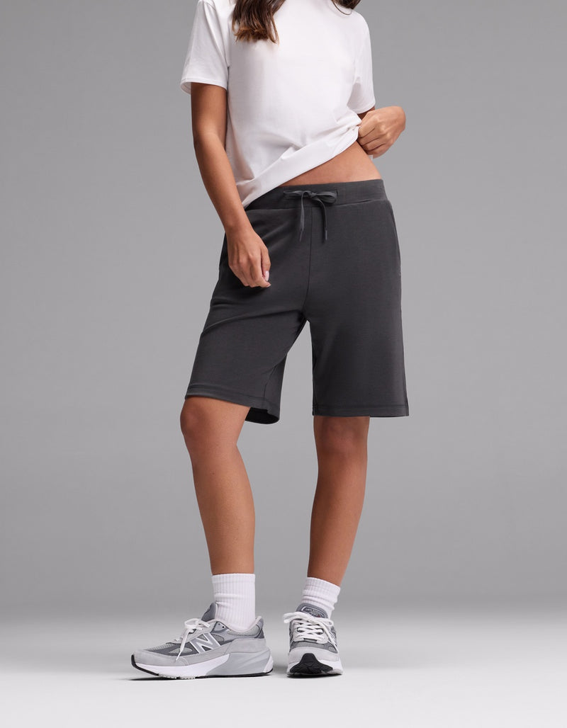 A person wearing a white t-shirt, Cozy Earth Women’s Brushed Bamboo Bermuda Shorts in dark gray, white socks, and gray sneakers stands against a plain gray background. Their face is not visible.