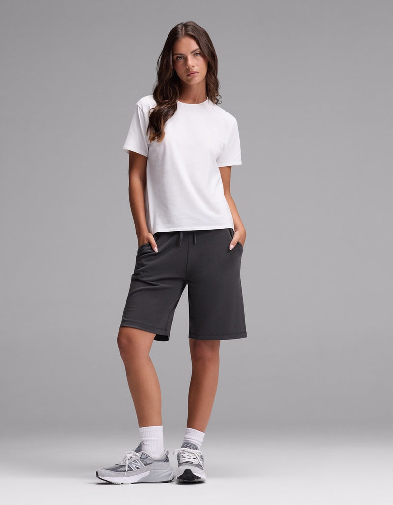 A woman stands against a gray background wearing Cozy Earth’s Women’s Brushed Bamboo Bermuda Shorts in dark gray, paired with a white t-shirt, white socks, and gray sneakers. Her hands are in her pockets as she looks slightly to the side.