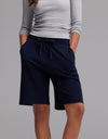 A person wearing a light gray long-sleeve shirt and Cozy Earth’s Women’s Brushed Bamboo Bermuda Shorts in dark navy, with hands in pockets, standing against a plain gray background.|Color:Navy