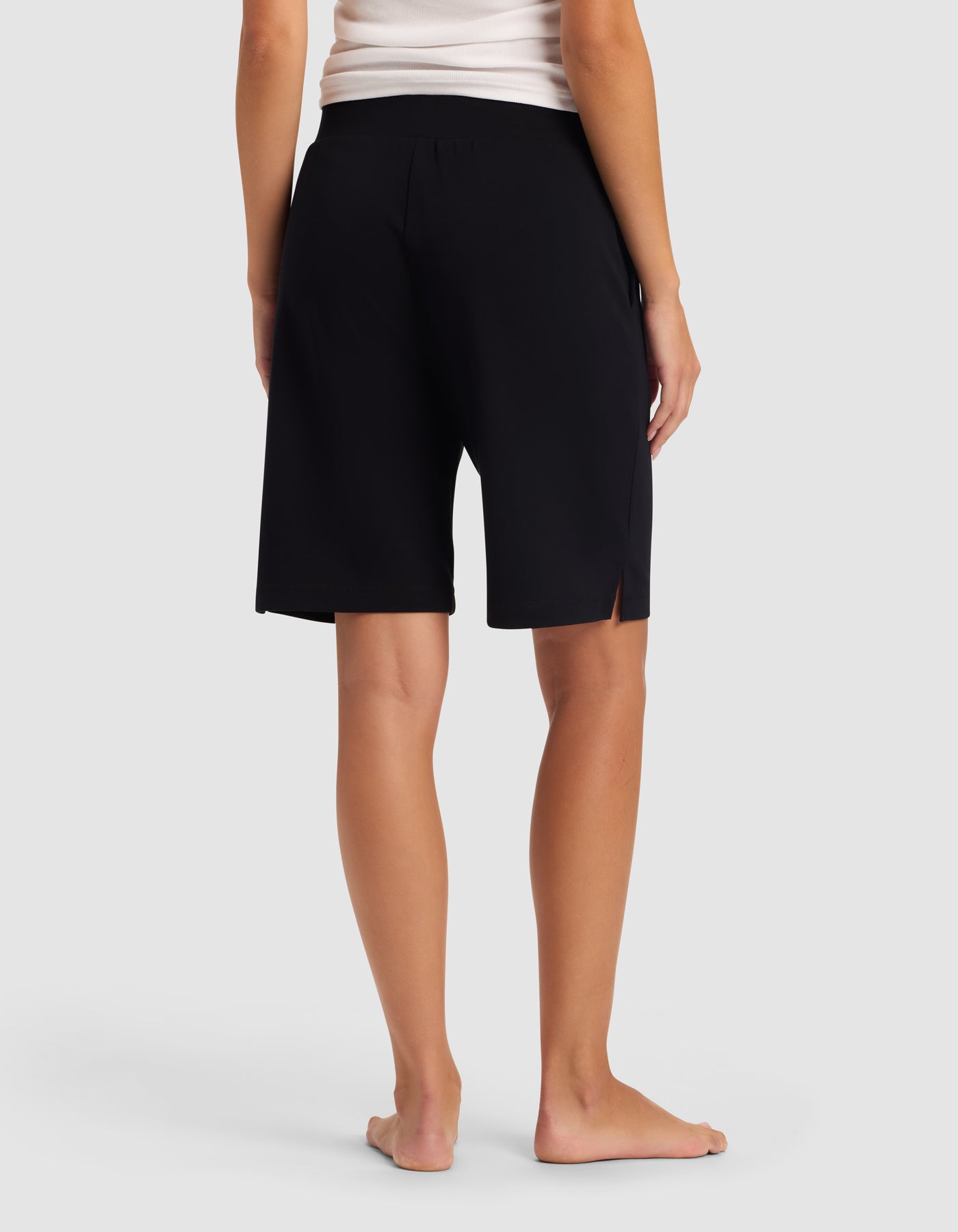 A woman stands barefoot on a white floor, seen from the back, wearing a white tank top and Cozy Earth Women's Brushed Bamboo Bermuda Shorts in black with a small side slit.|Color:Black