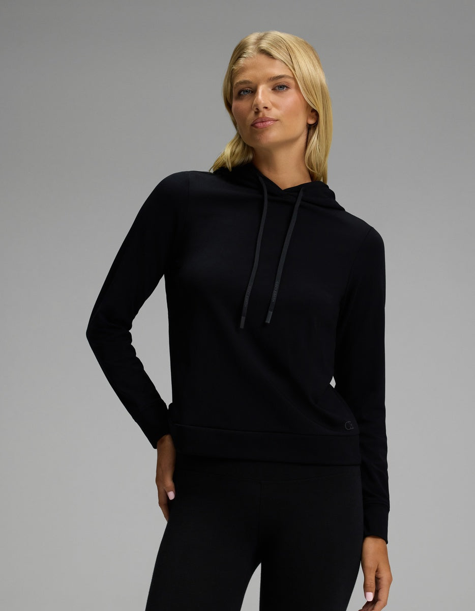A woman with long blonde hair wears the Cozy Earth Women’s Brushed Bamboo Hoodie in black, paired with black pants, standing against a plain gray background with a neutral expression.