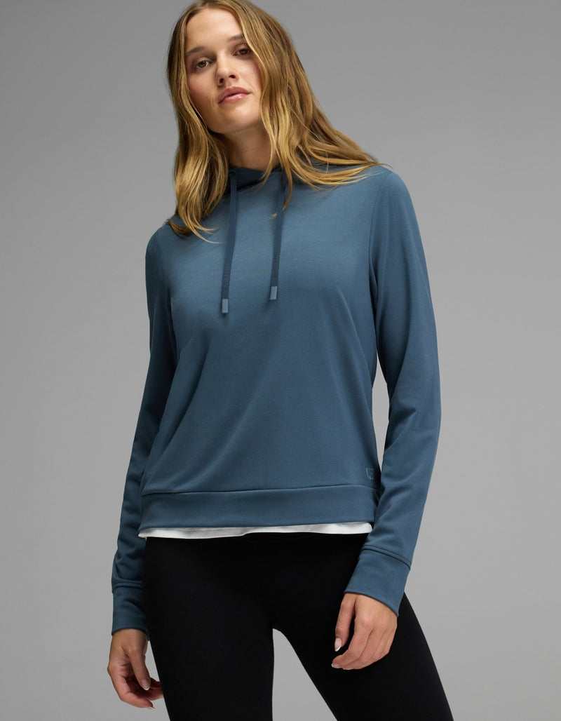 A woman with long, light brown hair wears the Cozy Earth Women’s Brushed Bamboo Hoodie in blue and black leggings, standing against a plain gray background.