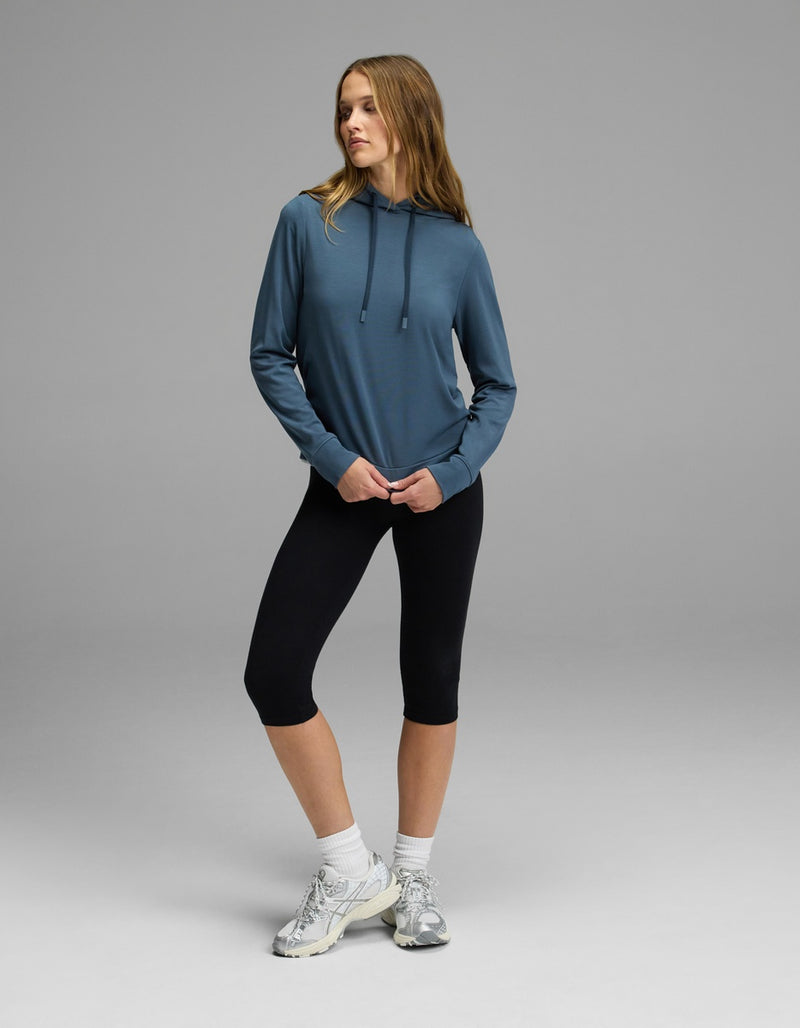 A woman stands on a gray background wearing the Cozy Earth Women’s Brushed Bamboo Hoodie, paired with black capri leggings, white socks, and white sneakers. She looks to the side with one hand in her hoodie pocket.