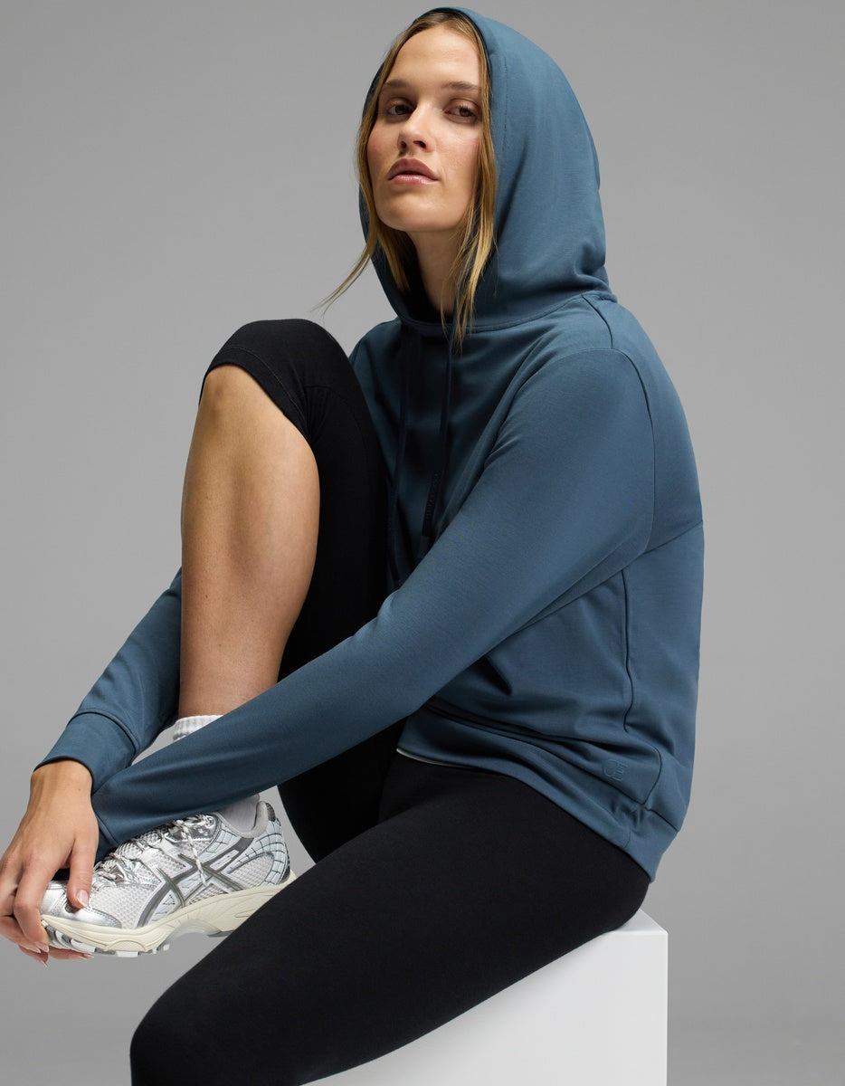 A woman wears the Cozy Earth Women’s Brushed Bamboo Hoodie, paired with black leggings and white sneakers, as she sits confidently on a white block against a gray background.