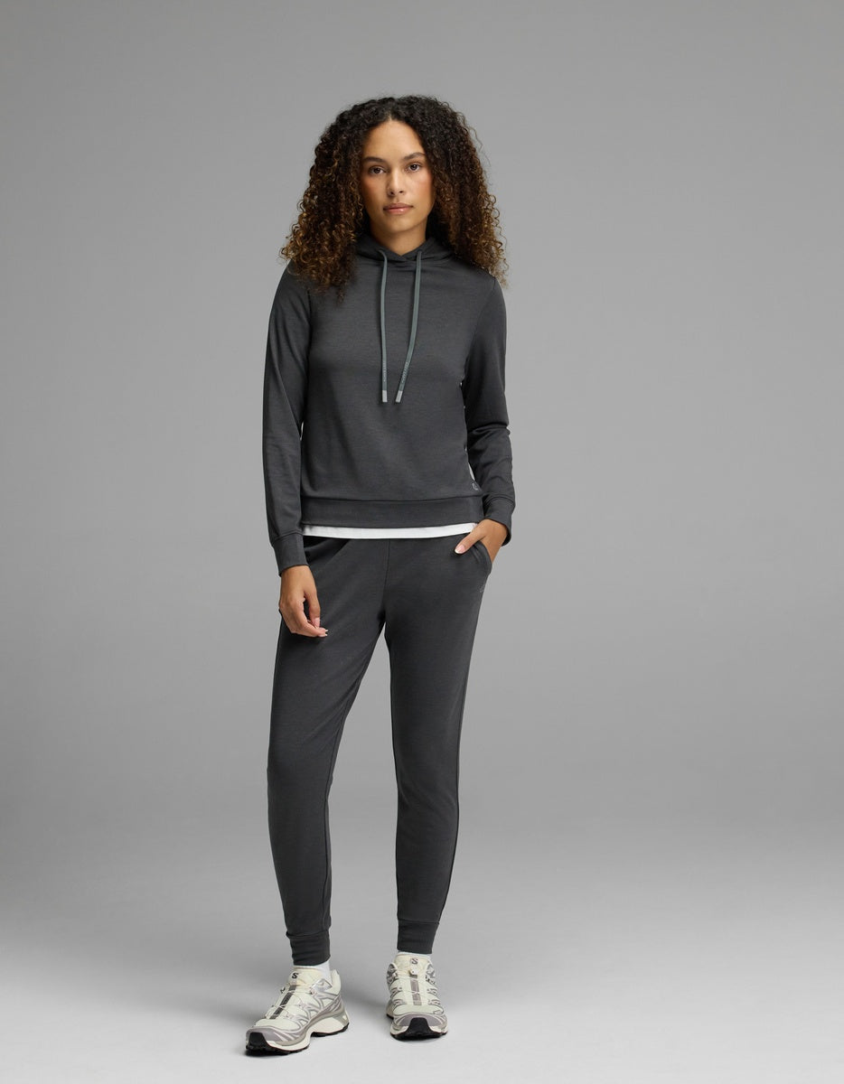 A woman with curly hair models the Cozy Earth Women’s Brushed Bamboo Hoodie & Jogger Set in a dark shade, paired with sneakers, standing against a plain gray background with a neutral expression.|Color:Charcoal