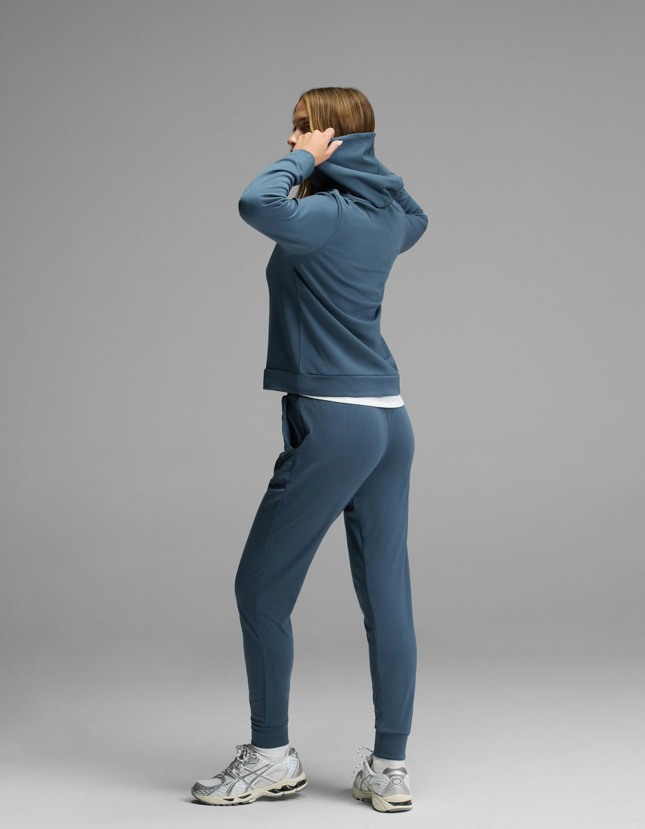 A woman wearing the Cozy Earth Women’s Brushed Bamboo Hoodie & Jogger Set stands on a gray background, facing away as she pulls up the hood over her head.|Color:Dawn