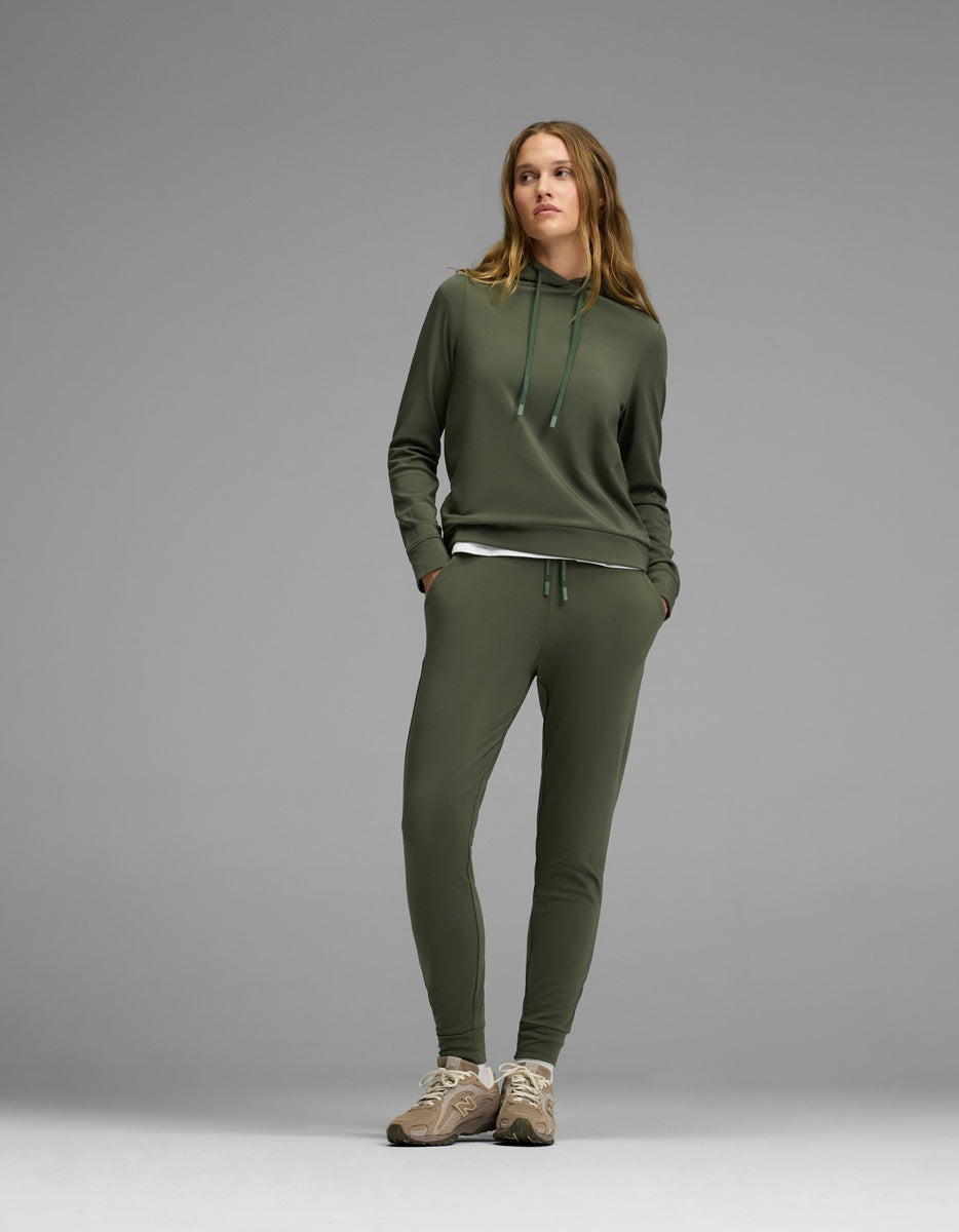 A woman stands against a gray background wearing the Cozy Earth Women’s Brushed Bamboo Hoodie & Jogger Set in olive green, paired with beige sneakers. Her hands are in her pockets as she looks slightly to the side.|Color:Olive