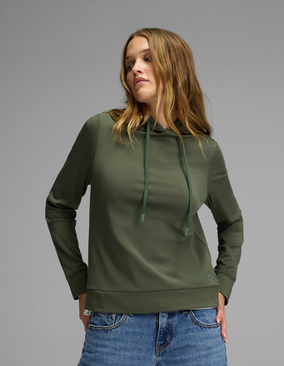 A woman wearing the Cozy Earth Women’s Brushed Bamboo Hoodie in green, paired with blue jeans, stands against a plain gray background, gazing slightly to the side with a relaxed expression.