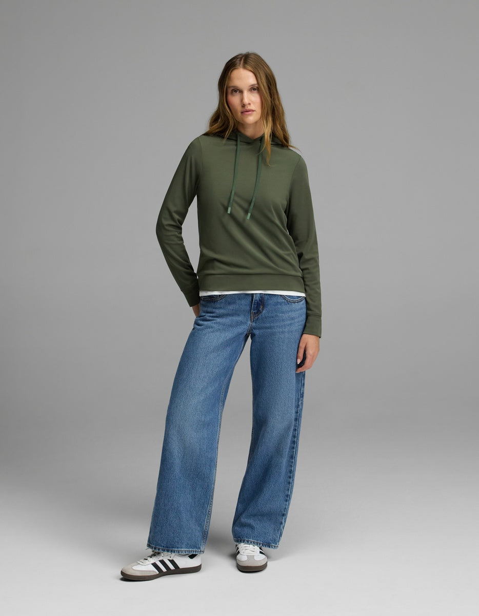 A woman stands facing forward against a plain gray background, wearing the Cozy Earth Women’s Brushed Bamboo Hoodie in green, paired with blue wide-leg jeans and white Adidas sneakers with black stripes.