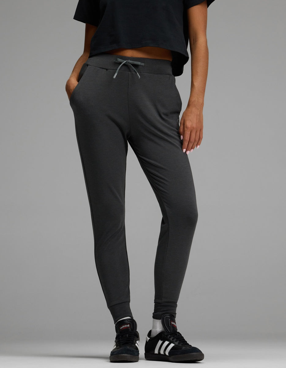 A person wearing Cozy Earth’s Women’s Brushed Bamboo Jogger in dark gray, paired with a black cropped shirt and black Adidas sneakers with white stripes, stands with hands in their pockets against a plain gray background.