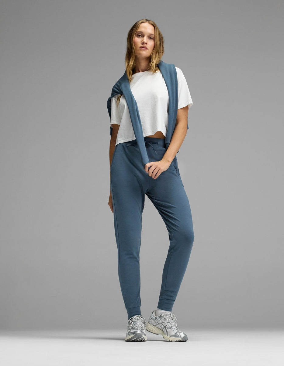 A person stands against a plain gray background in casual attire, featuring the Cozy Earth Women’s Brushed Bamboo Jogger in blue, a white cropped T-shirt, a matching blue sweatshirt draped over their shoulders, and silver sneakers.