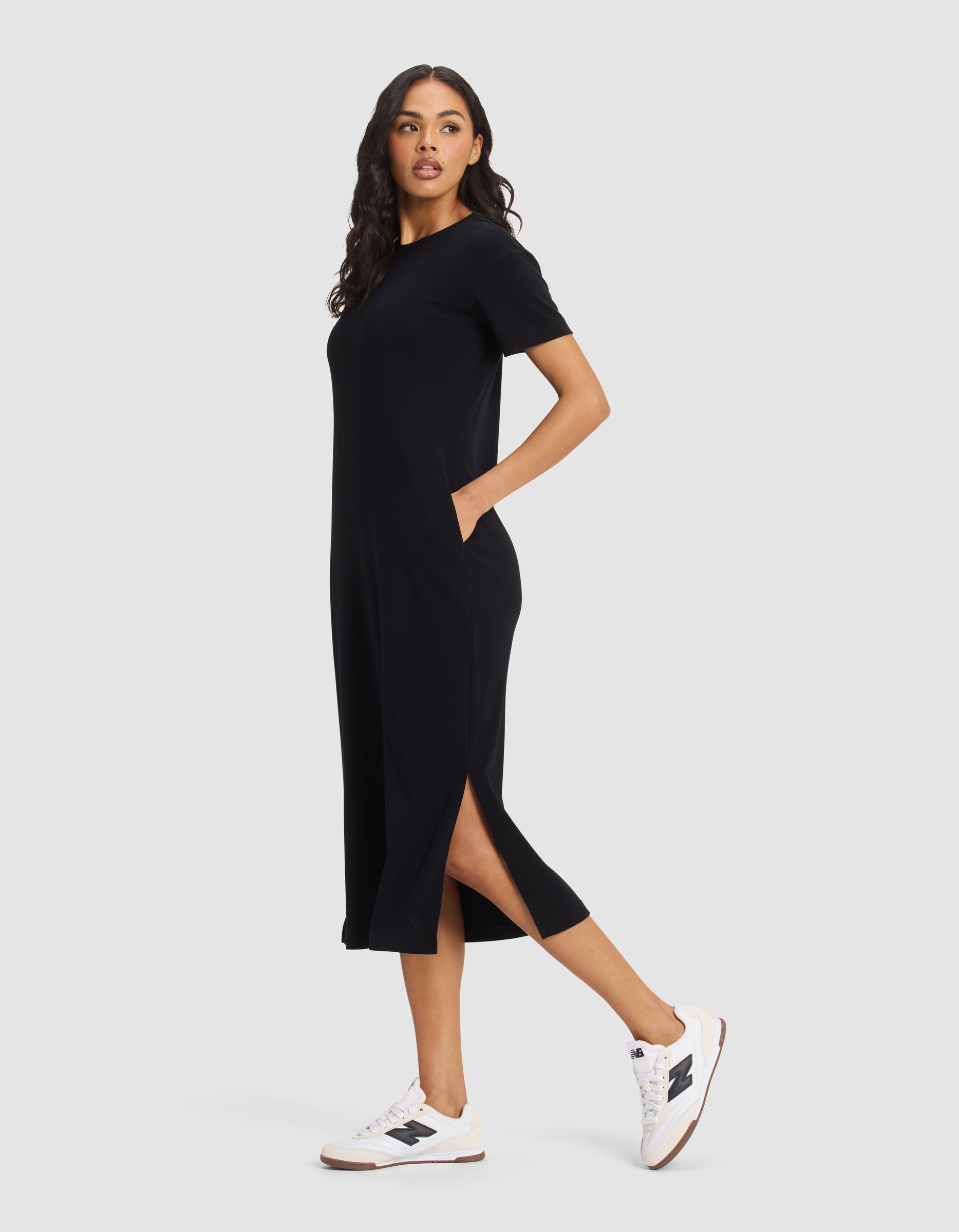 A woman stands sideways wearing the Cozy Earth Women's Brushed Bamboo Midi Dress with short sleeves, side slits, and pockets, paired with white sneakers. She looks ahead neutrally against a plain background.