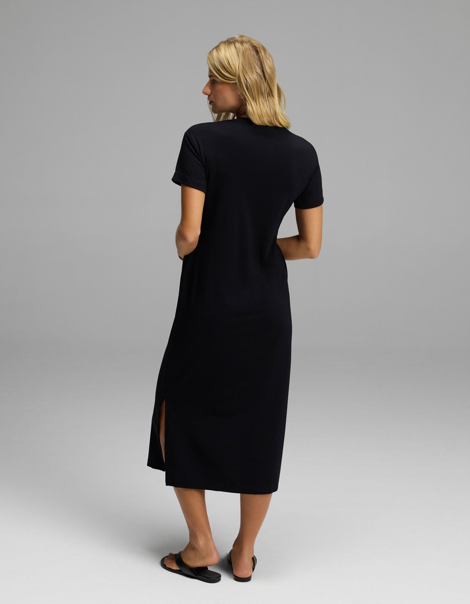 A woman with blonde hair stands facing away in a plain, light-colored studio, wearing Cozy Earth’s Women’s Brushed Bamboo Midi Dress—a black, short-sleeve style with a side slit—paired with black flats.