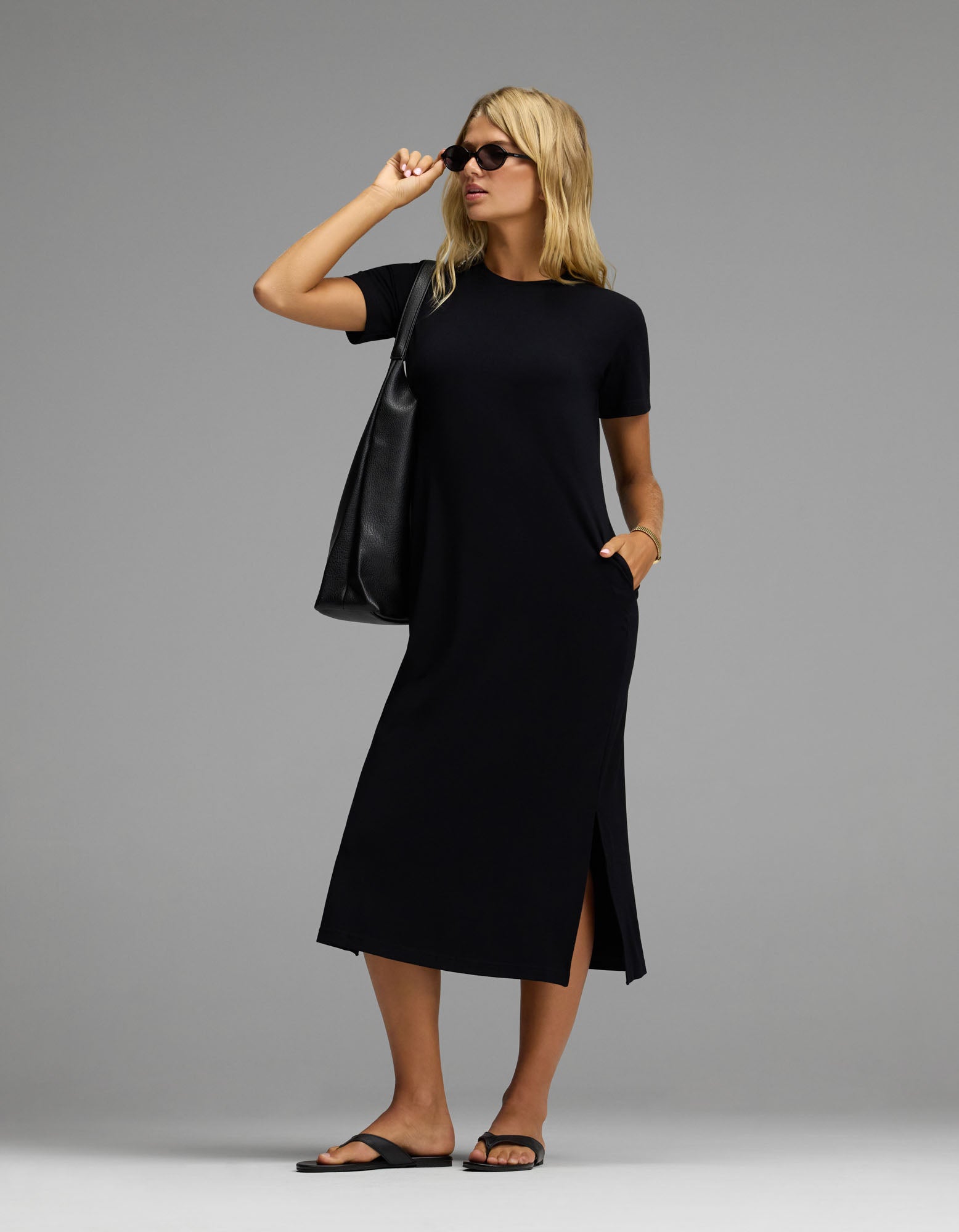 A woman stands confidently against a plain gray background, wearing the Cozy Earth Women’s Brushed Bamboo Midi Dress in black with short sleeves and side slits. She completes her look with black sandals, sunglasses, and a large black tote bag.