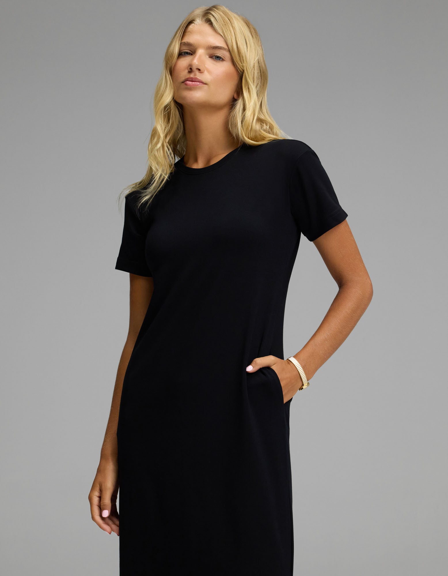 A woman with long blonde hair wears the Cozy Earth Women’s Brushed Bamboo Midi Dress in black, paired with a bracelet, standing against a plain gray background with one hand in her pocket.
