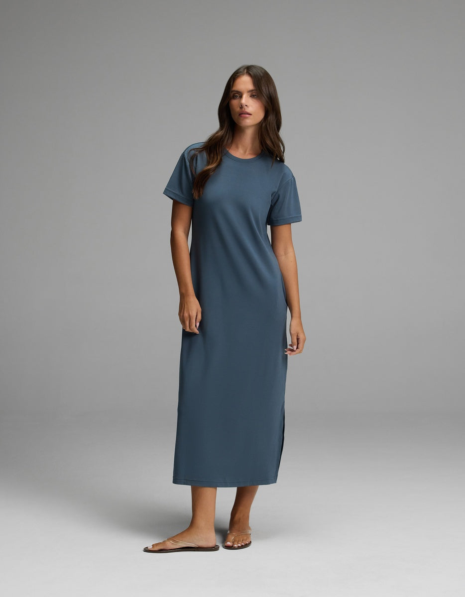 A woman stands in a studio against a plain gray background, wearing Cozy Earth’s Women’s Brushed Bamboo Midi Dress in Dawn—a loose, short-sleeved, ankle-length style—paired with sandals.|Color:Dawn