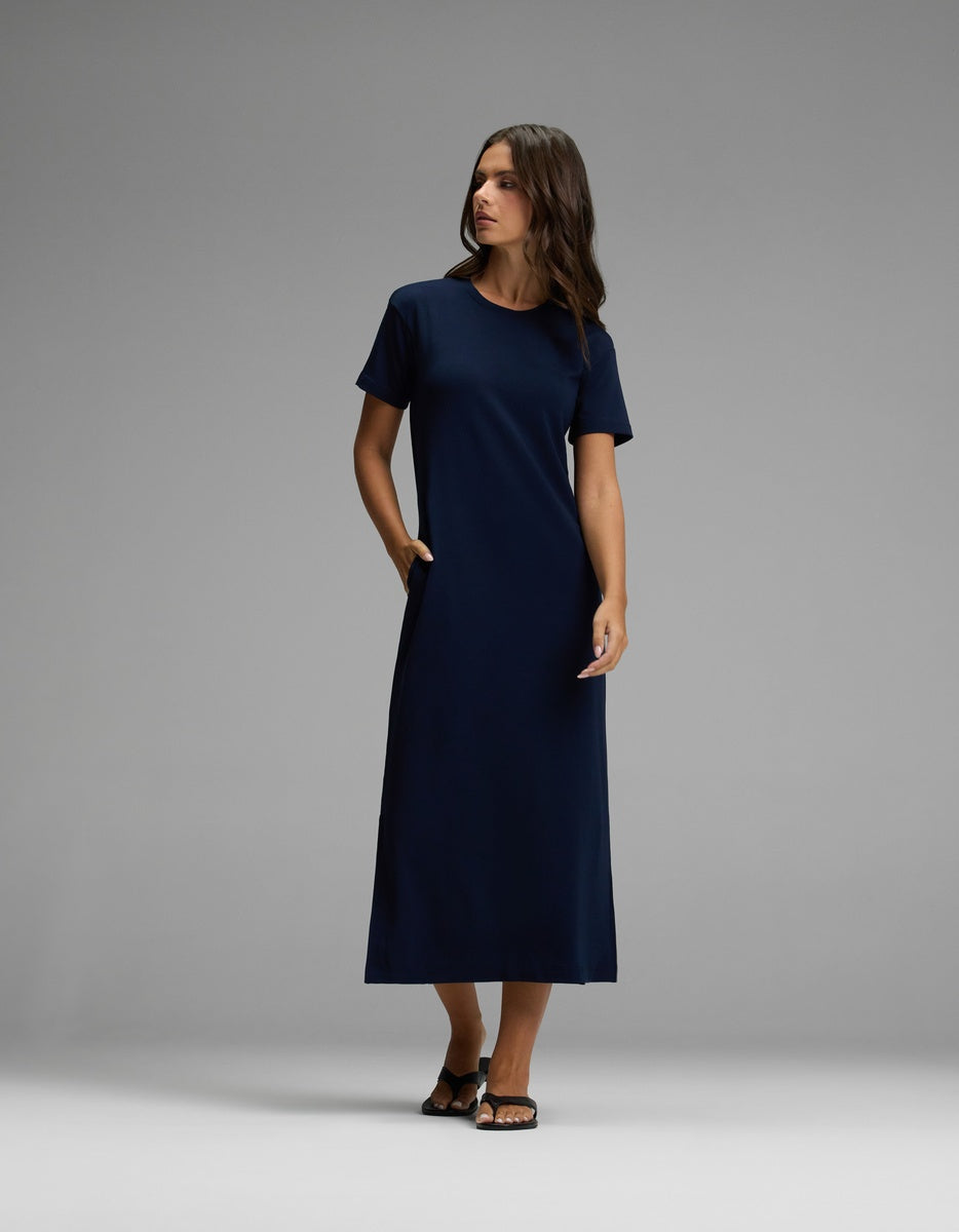 A woman stands in a studio against a plain gray background, wearing the Cozy Earth Women’s Brushed Bamboo Midi Dress in navy. The dress features short sleeves, an ankle-length hem, and convenient pockets. She has dark hair, wears sandals, and gazes slightly to the side.|Color:Navy