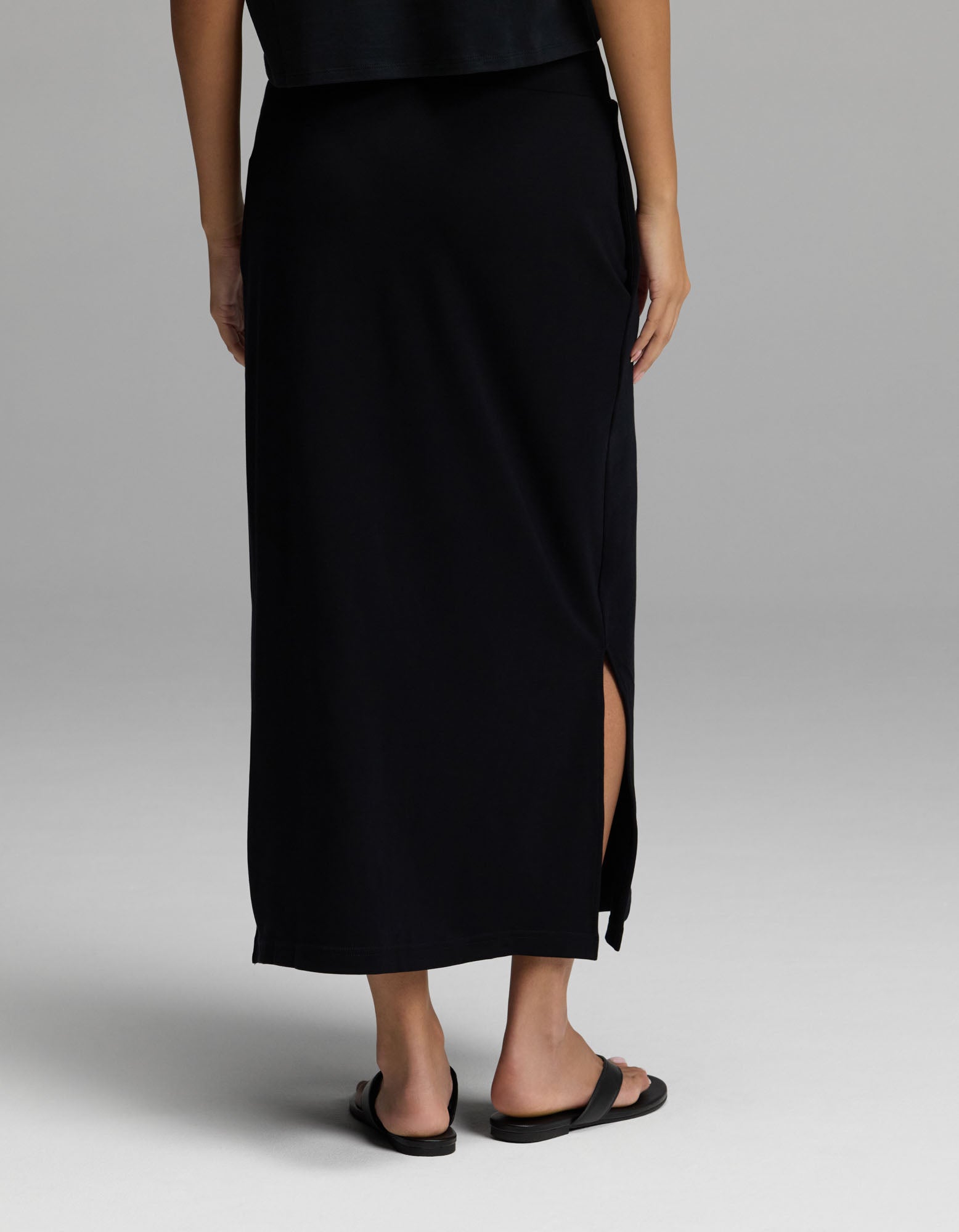 A person is shown from the back, wearing Cozy Earth’s Women’s Brushed Bamboo Midi Skirt in black—with a side slit—paired with black sandals, standing against a light gray background. Their upper body is partially visible.