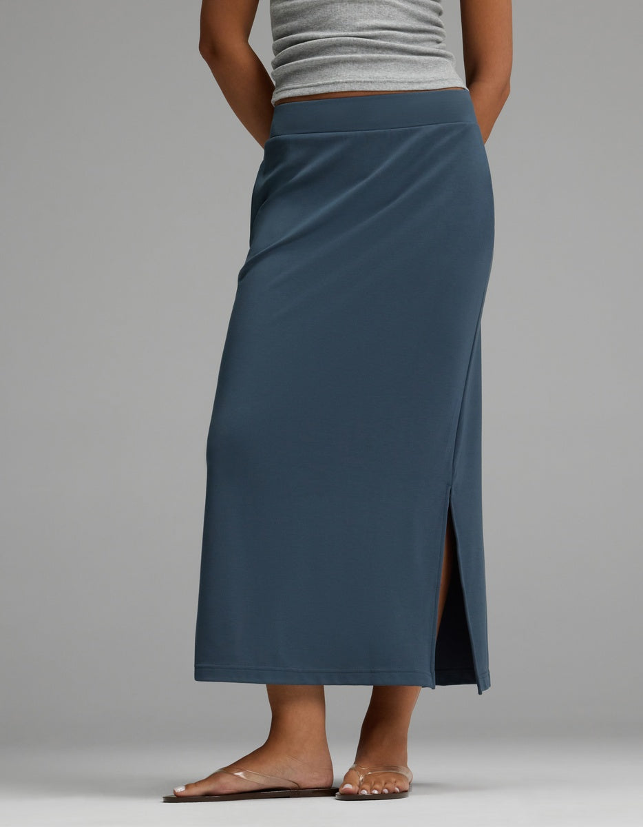 A person is shown from the waist down, barefoot, wearing a Cozy Earth Women’s Brushed Bamboo Midi Skirt in blue with a side slit, paired with a gray tank top, standing against a light gray background.|Color:Dawn