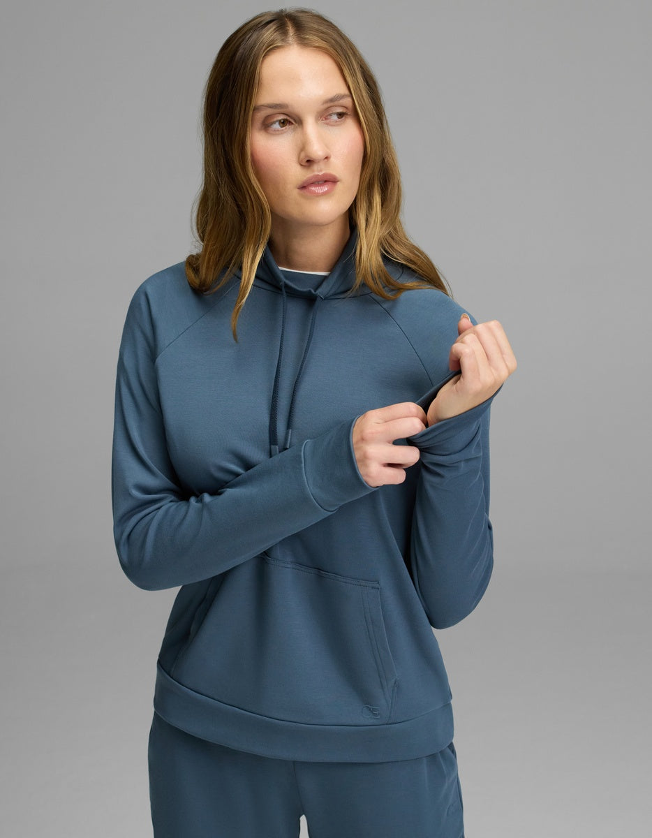 A woman wearing the Cozy Earth Women’s Brushed Bamboo Mock Neck in Dawn stands against a gray background, looking to the side while pulling at the sleeve’s thumb hole.