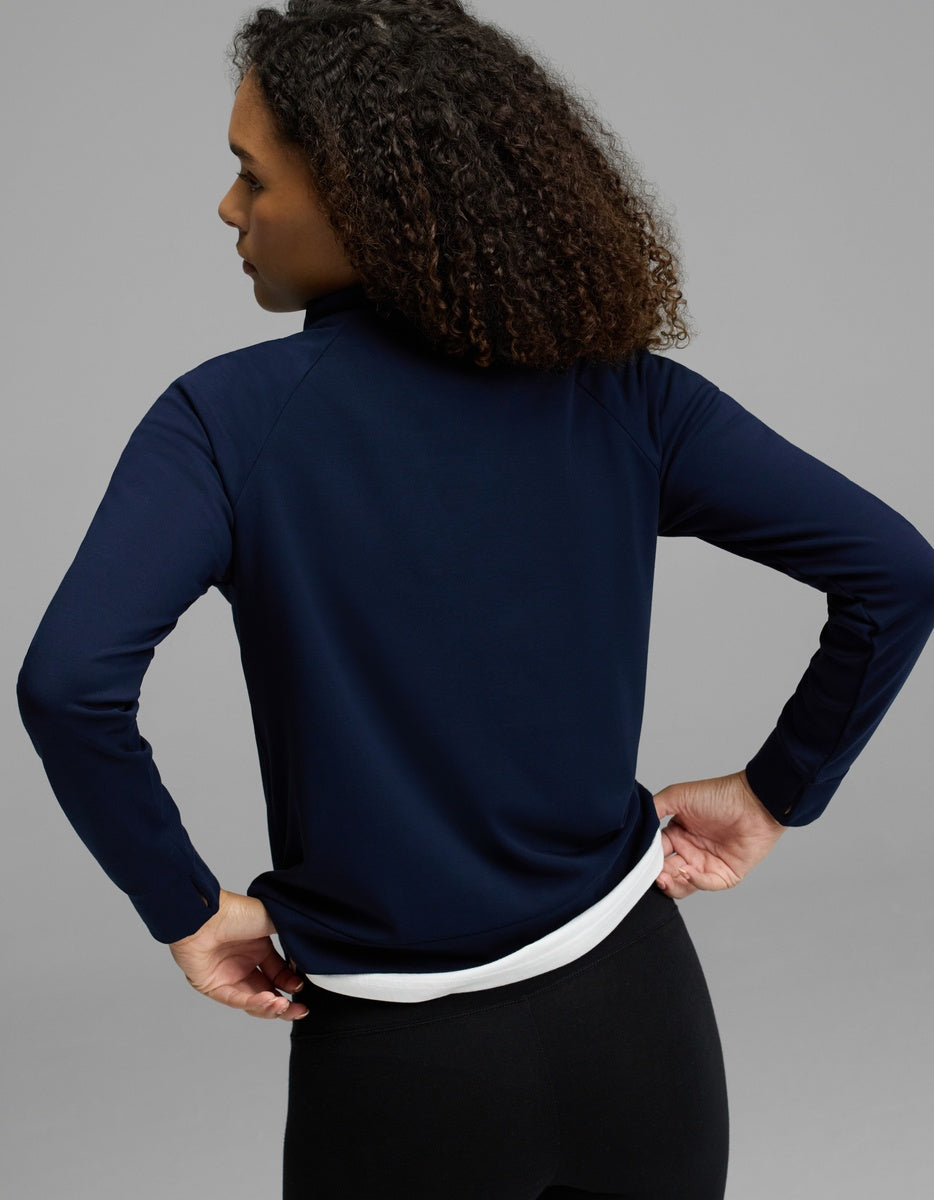 A woman with curly hair, wearing the Cozy Earth Women’s Brushed Bamboo Mock Neck in navy over a white shirt and black pants, stands with her back to the camera against a plain gray background.|Color:Navy