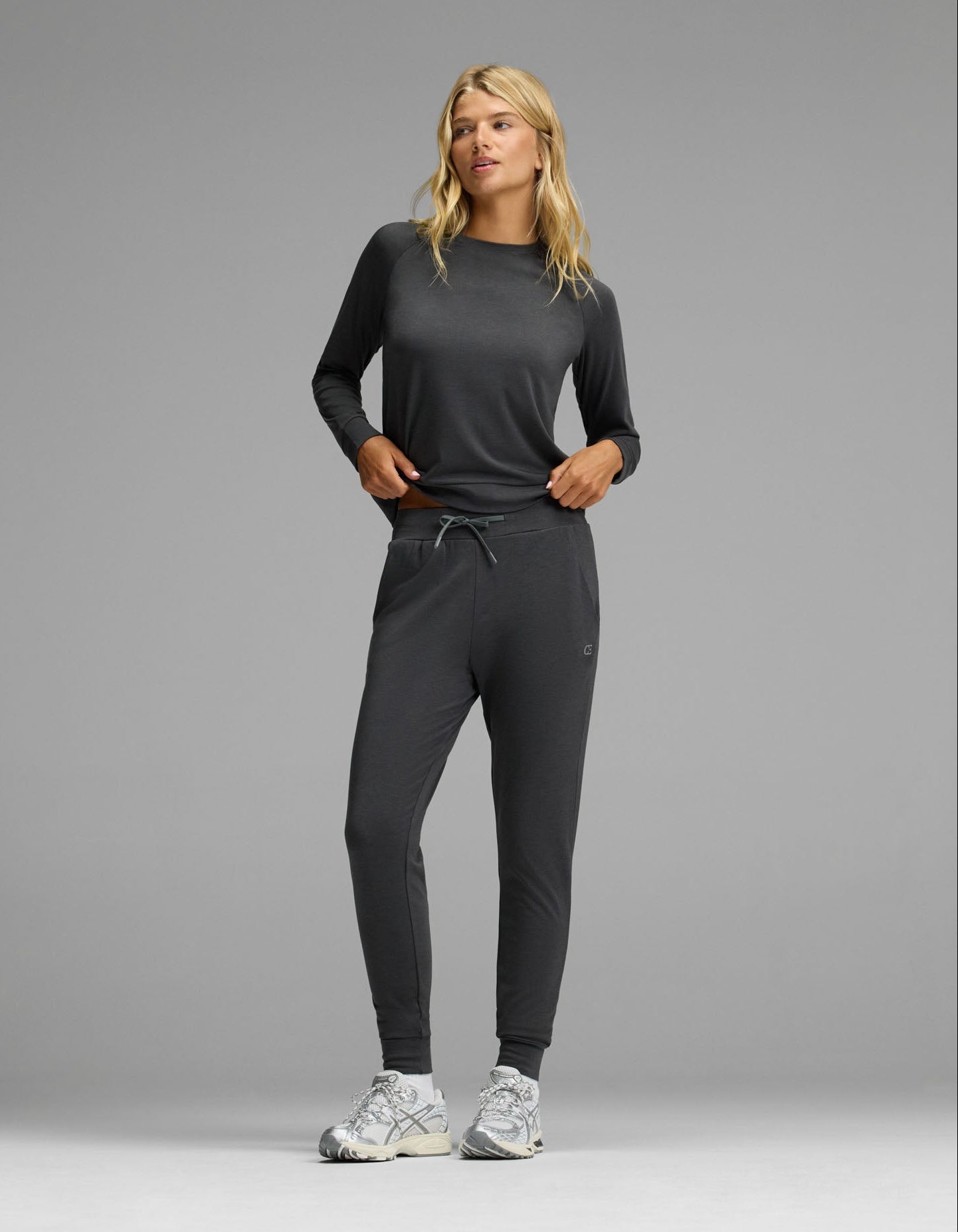 A woman with blonde hair stands against a gray background wearing the Cozy Earth Women’s Brushed Bamboo Jogger Set in Charcoal, paired with white sneakers. Her hands are in her pockets.