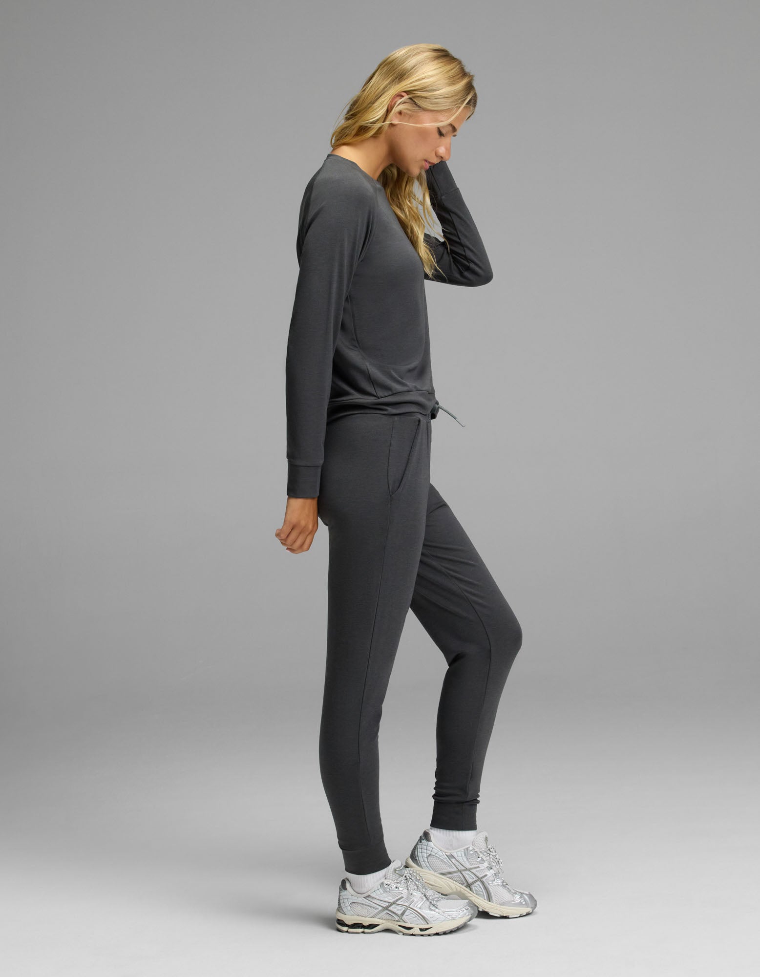 A woman with long blonde hair is shown in profile against a plain gray background, wearing the Cozy Earth Women’s Brushed Bamboo Jogger Set—a dark gray long-sleeve top and matching jogger pants—paired with white athletic shoes.