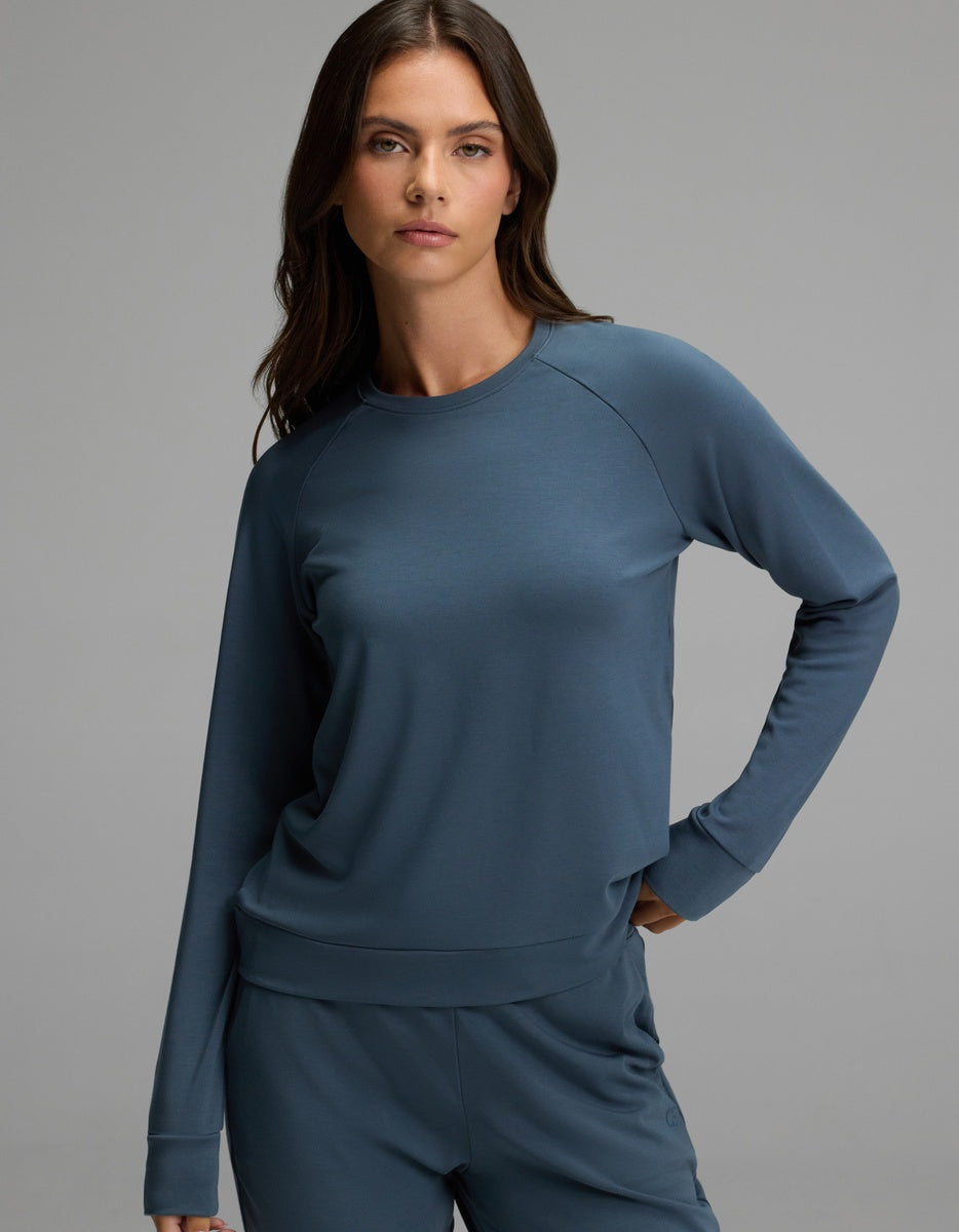 A woman with long brown hair wears the Cozy Earth Women’s Brushed Bamboo Pullover Crew in blue, standing against a plain grey background with a neutral expression.