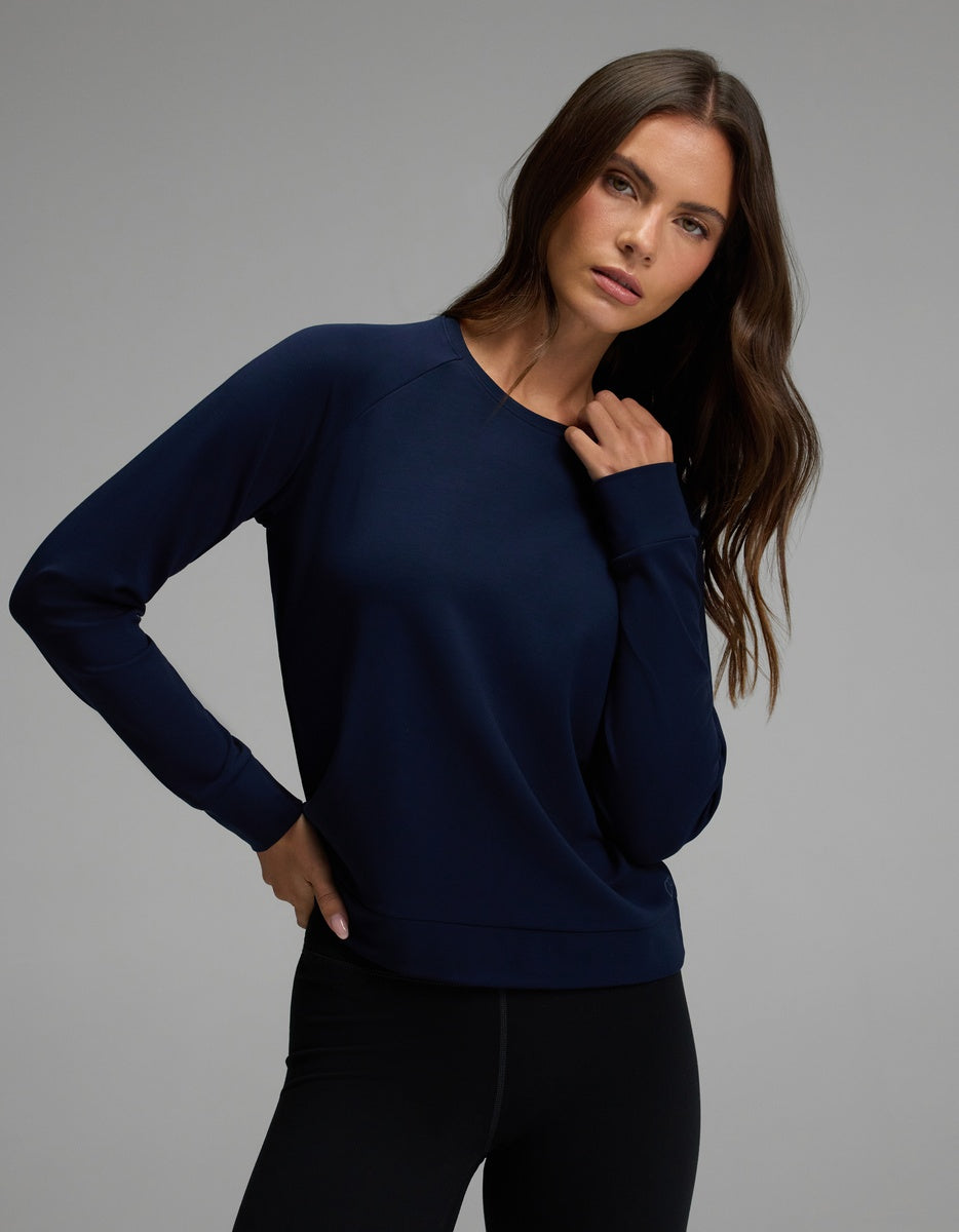 A woman with long brown hair wears the Cozy Earth Women’s Brushed Bamboo Pullover Crew in navy blue, paired with black leggings, standing against a plain gray background.