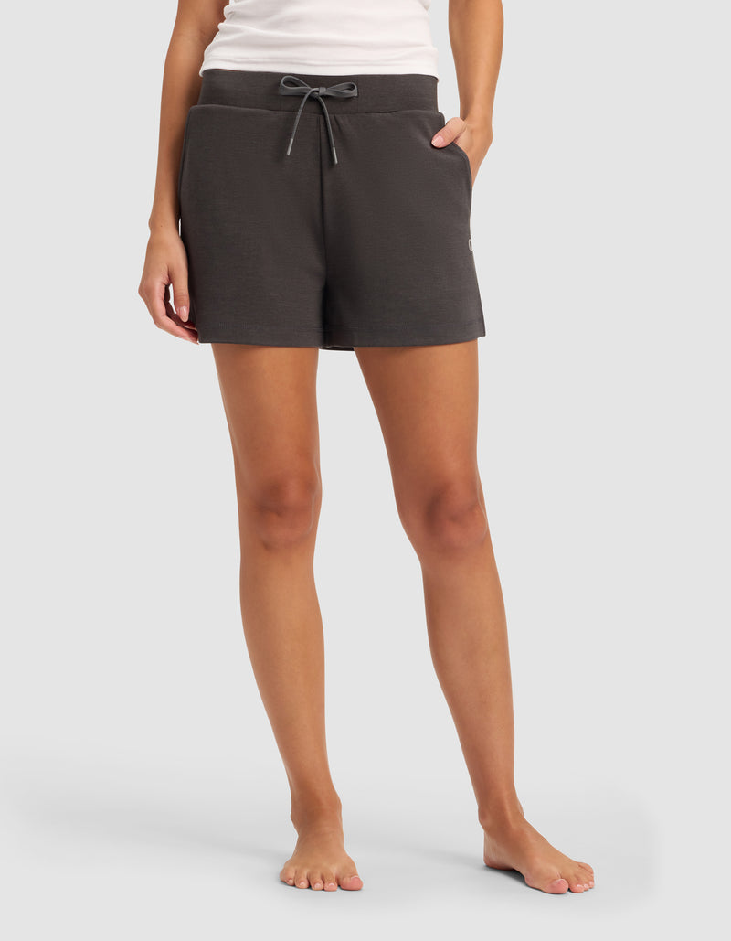 A woman stands barefoot against a plain light gray background, wearing Cozy Earth’s Women’s Brushed Bamboo Shorts in dark gray along with a white tank top.