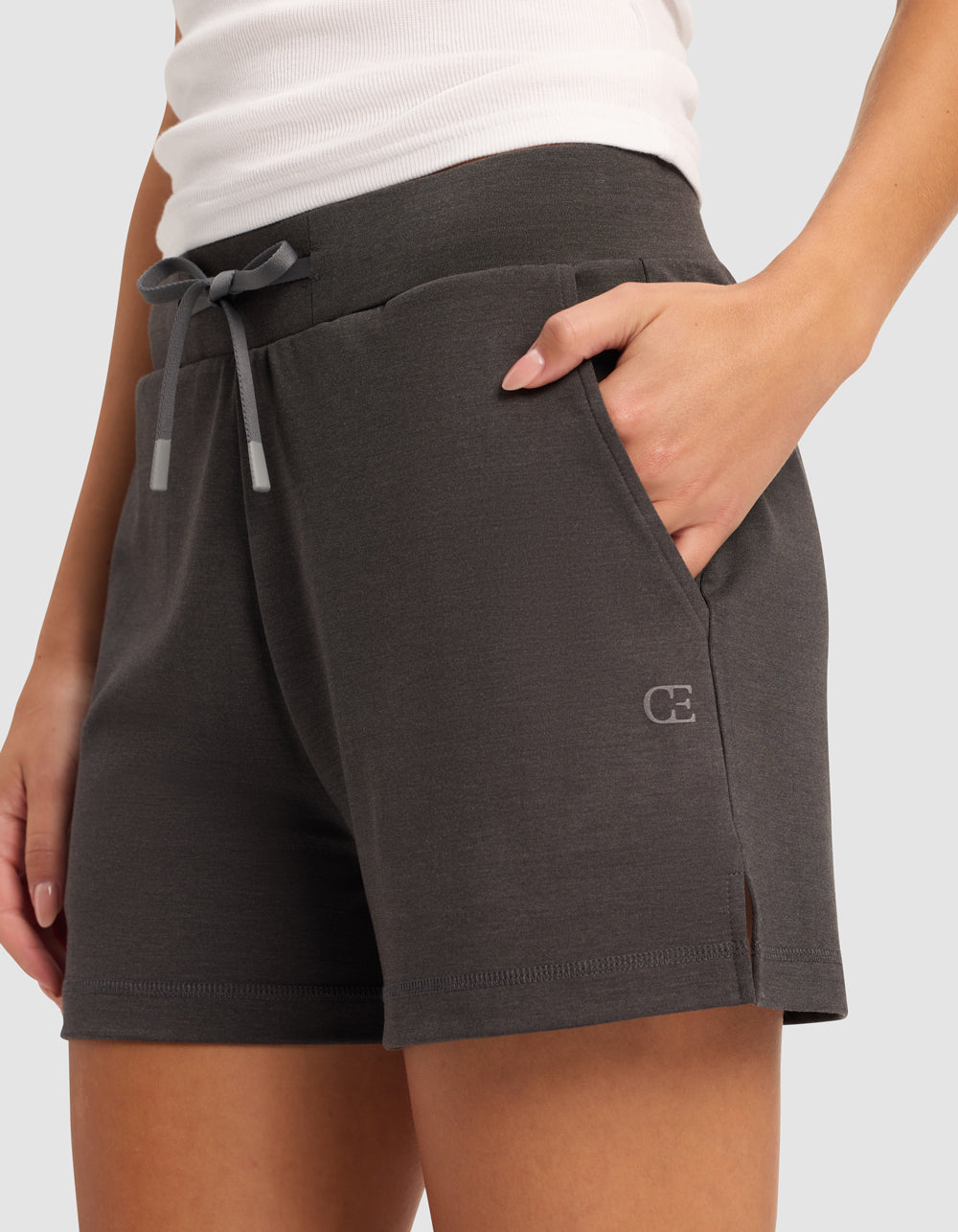 A person wears Cozy Earth Women’s Brushed Bamboo Shorts in dark gray, featuring a small left-leg logo and pockets, paired with a white tank top. Their left hand is tucked into the shorts’ pocket.