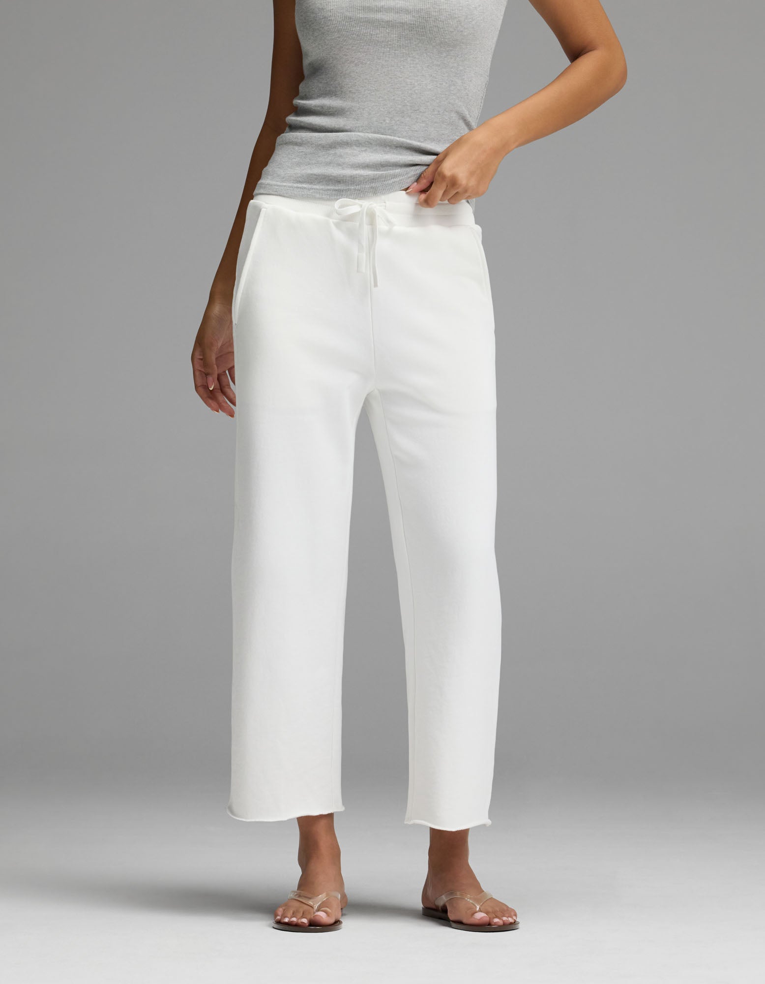 A person wearing the Cozy Earth Women’s Chelsea Cropped Pant in Bone stands on a gray background, with one hand adjusting the waistband. Their face is not visible.|Color:Bone