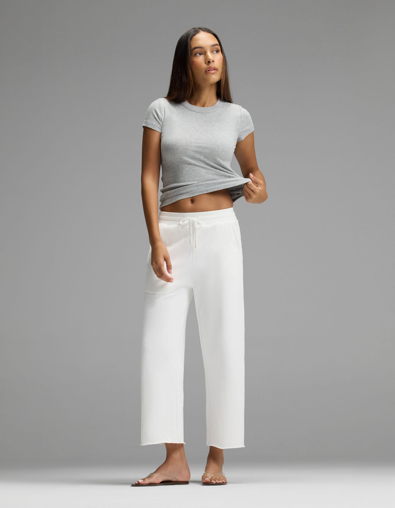 A woman wears the Cozy Earth Women’s Chelsea Cropped Pant, standing barefoot on a gray background in a gray t-shirt, lifting one side of her shirt and gazing slightly upward.