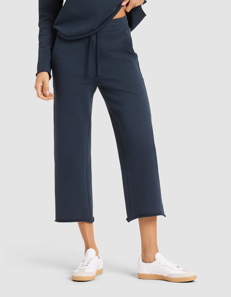 A woman stands against a plain light gray background wearing Cozy Earth's Women's Chelsea Cropped Pant in navy blue with a matching top and white sneakers.