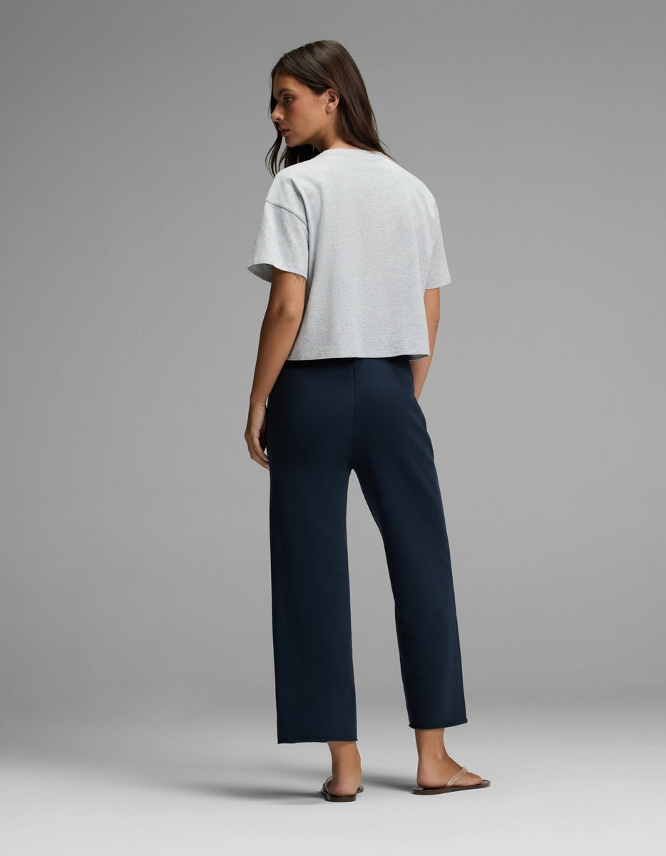A woman stands facing away against a plain gray background, wearing Cozy Earth’s Women’s Chelsea Cropped Pant in dark blue, paired with a light gray short-sleeve top and brown sandals.|Color:Eclipse