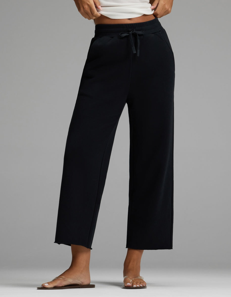 A person is wearing the Cozy Earth Women’s Chelsea Cropped Pant in Jet Black, styled with a white cropped tank top and clear flat sandals. The image highlights the legs and lower torso against a plain gray background.
