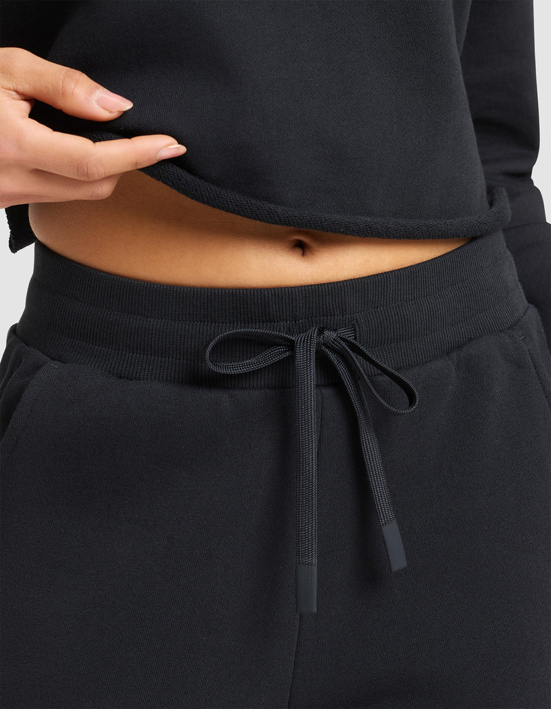 A woman models the Cozy Earth Women's Chelsea Cropped Pant in black, paired with a matching top that's lifted to reveal her bare midriff and navel.