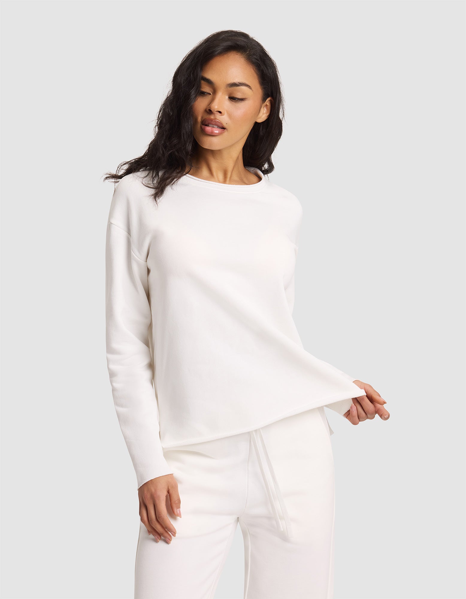 A woman poses against a light gray background wearing the Cozy Earth Women's Chelsea Pullover and matching pants in all white, holding the hem of the top with one hand while looking slightly downward. 