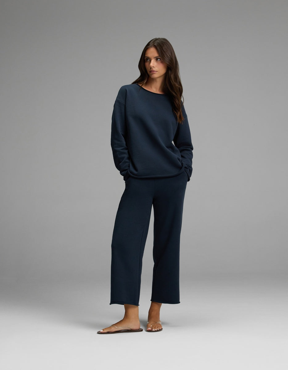 A woman stands in a relaxed pose, hands in her pockets, wearing the Cozy Earth Women’s Chelsea Cropped Set—a matching navy blue long-sleeve top and wide-leg pants—with brown sandals, against a plain gray background.