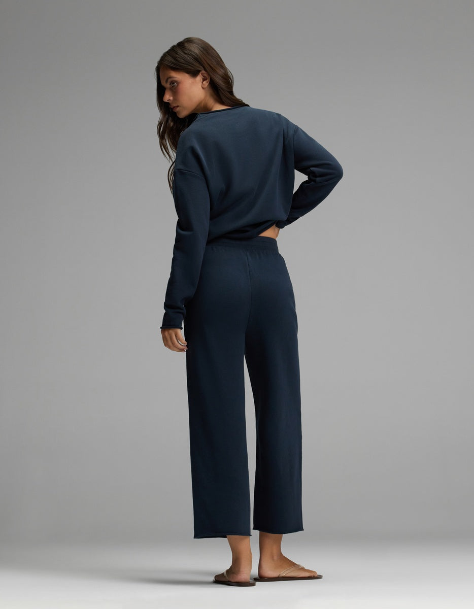 A woman with long brown hair stands barefoot on a light gray floor against a plain gray background, facing away and looking over her left shoulder. She is wearing the Cozy Earth Women’s Chelsea Cropped Set in dark blue, featuring a long-sleeve top and matching pants.|Color:Eclipse